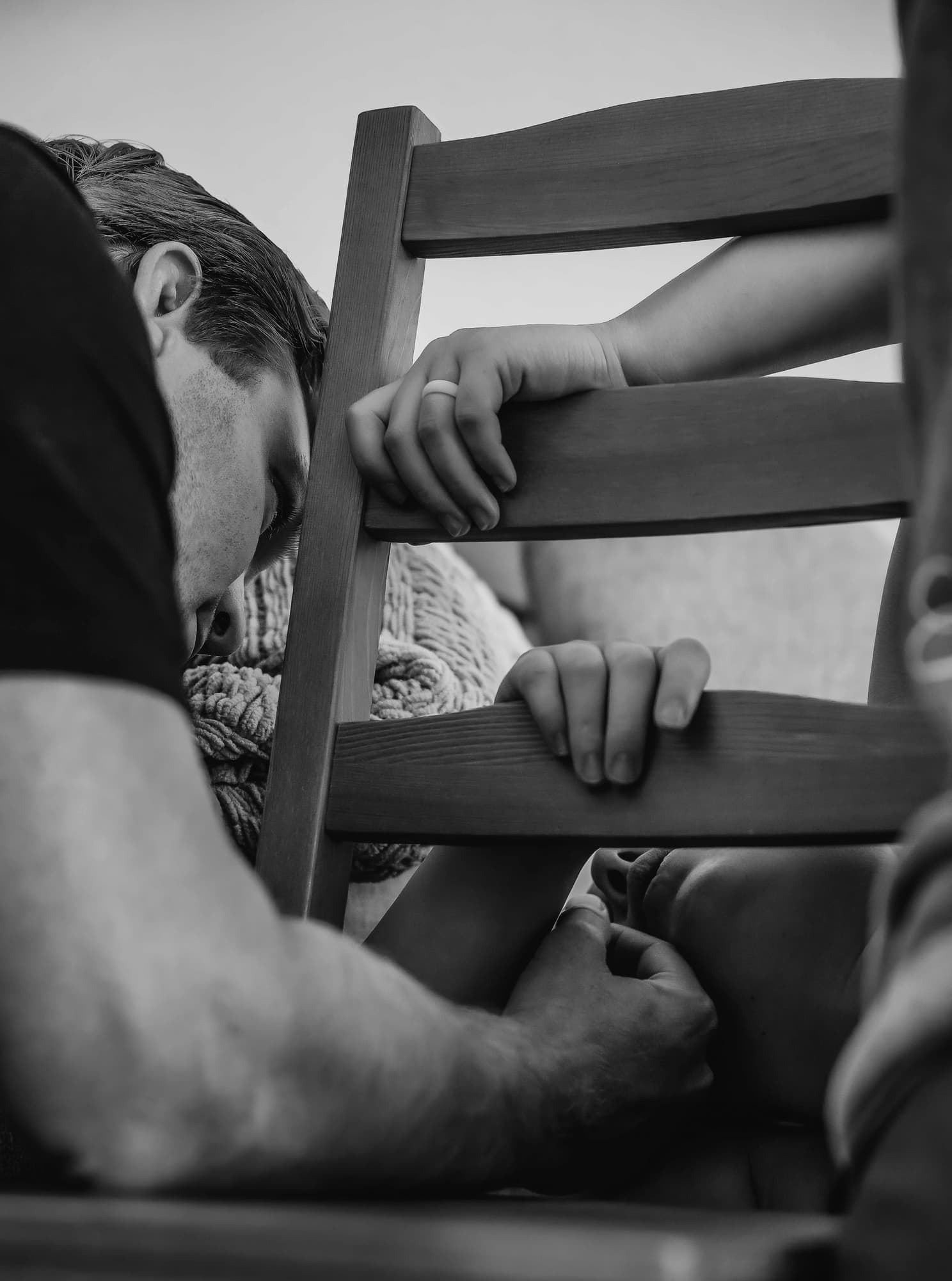 Black and white photo of a laboring mother draped over a wooden chair, her partner resting close