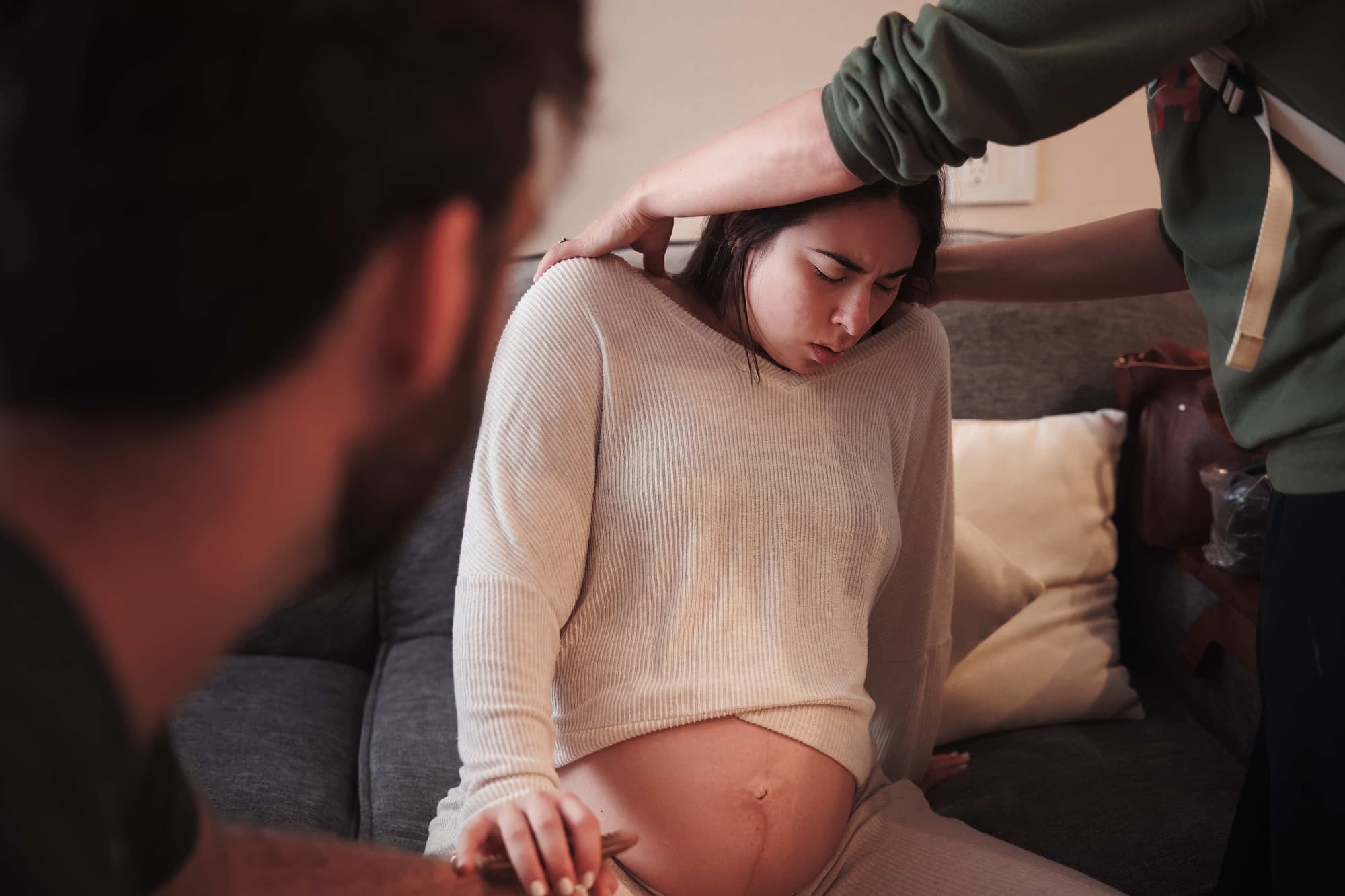 A laboring mother on a couch, eyes closed, as a support person rests a hand on her head and shoulder